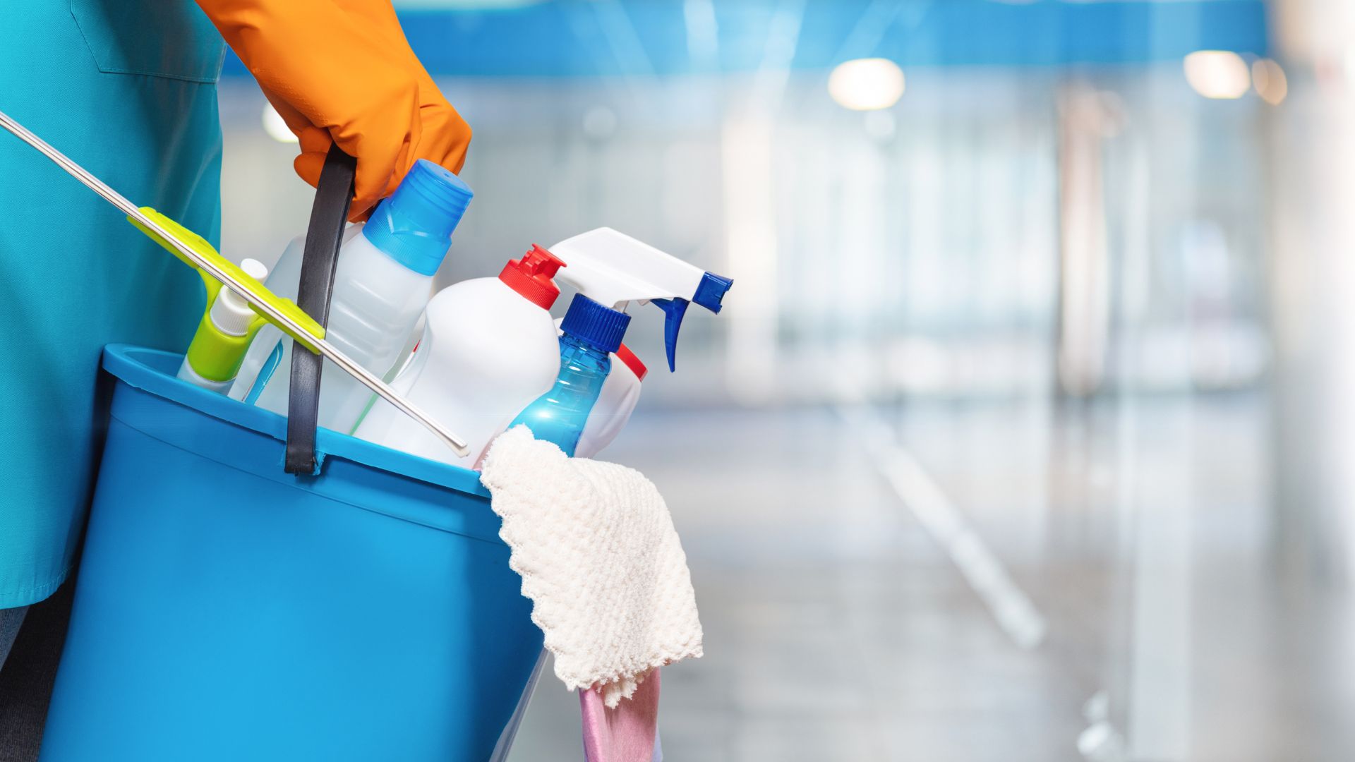 Cleaning supplies in blue bucket with gloved hand, spray bottles, and towel